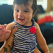 Alicia is registered to the contest to win money with this photo: baby, child, infant, smiling, happy, portrait, sitting, striped_outfit, red_flower, indoors, family, adult, lap, tattoo, hands, face, hair, couch, window, natural_light