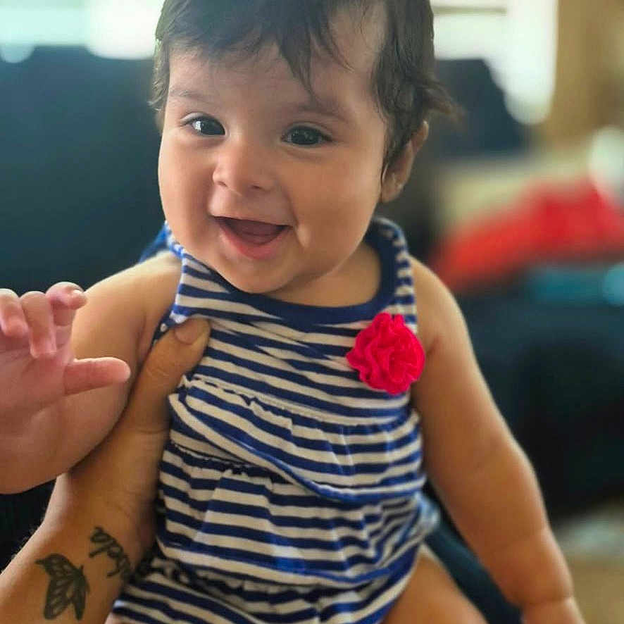 Alicia is registered to the contest to win money with this photo: baby, child, infant, smiling, happy, portrait, sitting, striped_outfit, red_flower, indoors, family, adult, lap, tattoo, hands, face, hair, couch, window, natural_light