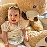 baby, child, cozy, cute, floor, hair_bows, happy, home, indoor, infant, plush, portrait, sitting, smiling, soft_toys, stuffed_animal, sunlight, teddy_bear, white_clothing, window
