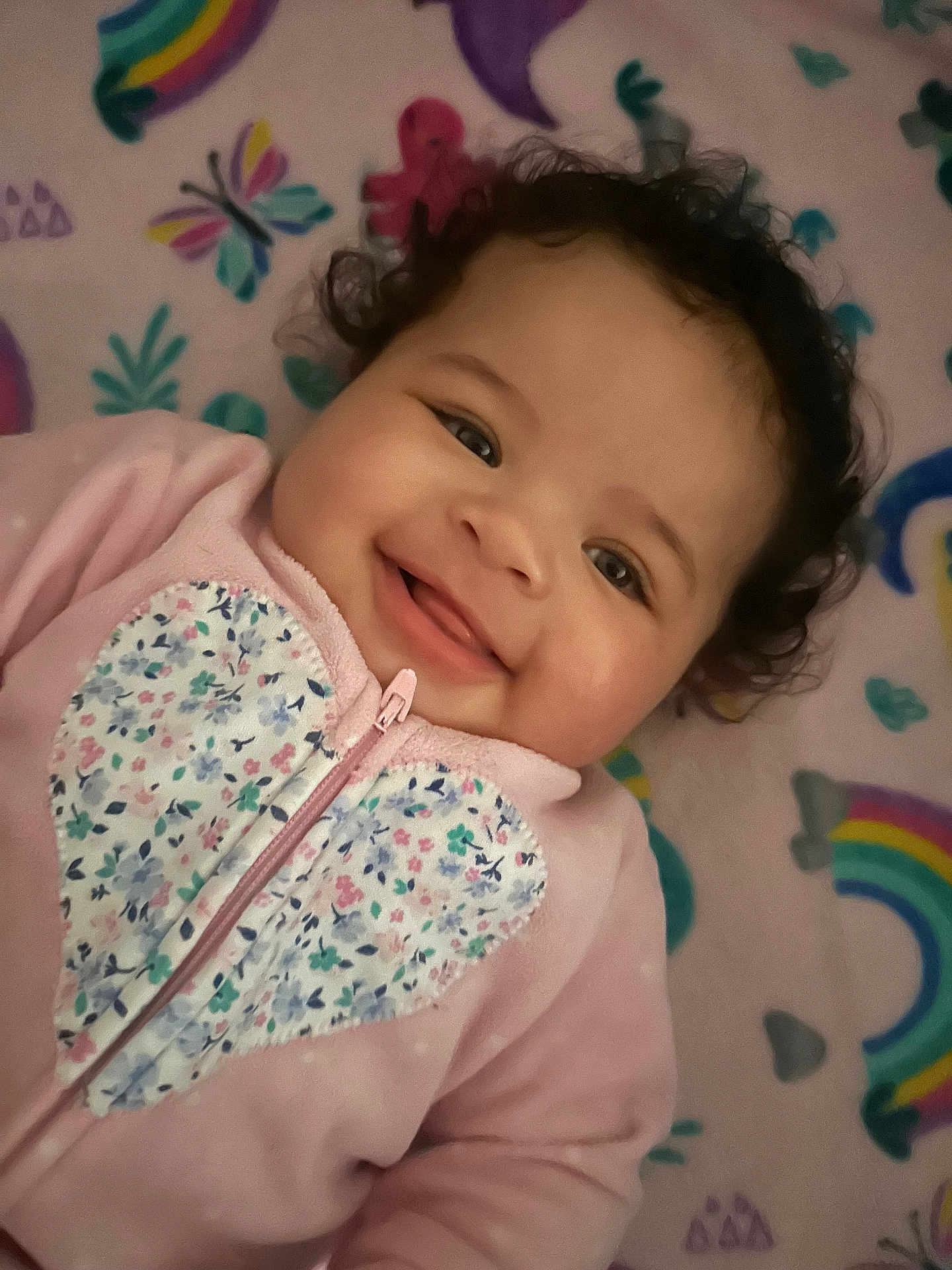 Kyla is registered to the contest to win money with this photo: baby, smiling, curly_hair, pink_clothing, onesie, floral_pattern, heart_shape, blanket, rainbow, butterfly, colorful, infant, cute, happy, child, playful, portrait, lying_down, soft_texture, young