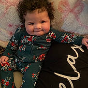 Kyla is registered to the contest to win money with this photo: baby, child, curly_hair, onesie, floral_pattern, blanket, cushion, smile, cheeks, indoors, portrait, cute, infant, cozy, relaxed, person, seated, playful, young_child, expression