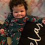 baby, child, curly_hair, onesie, floral_pattern, blanket, cushion, smile, cheeks, indoors, portrait, cute, infant, cozy, relaxed, person, seated, playful, young_child, expression