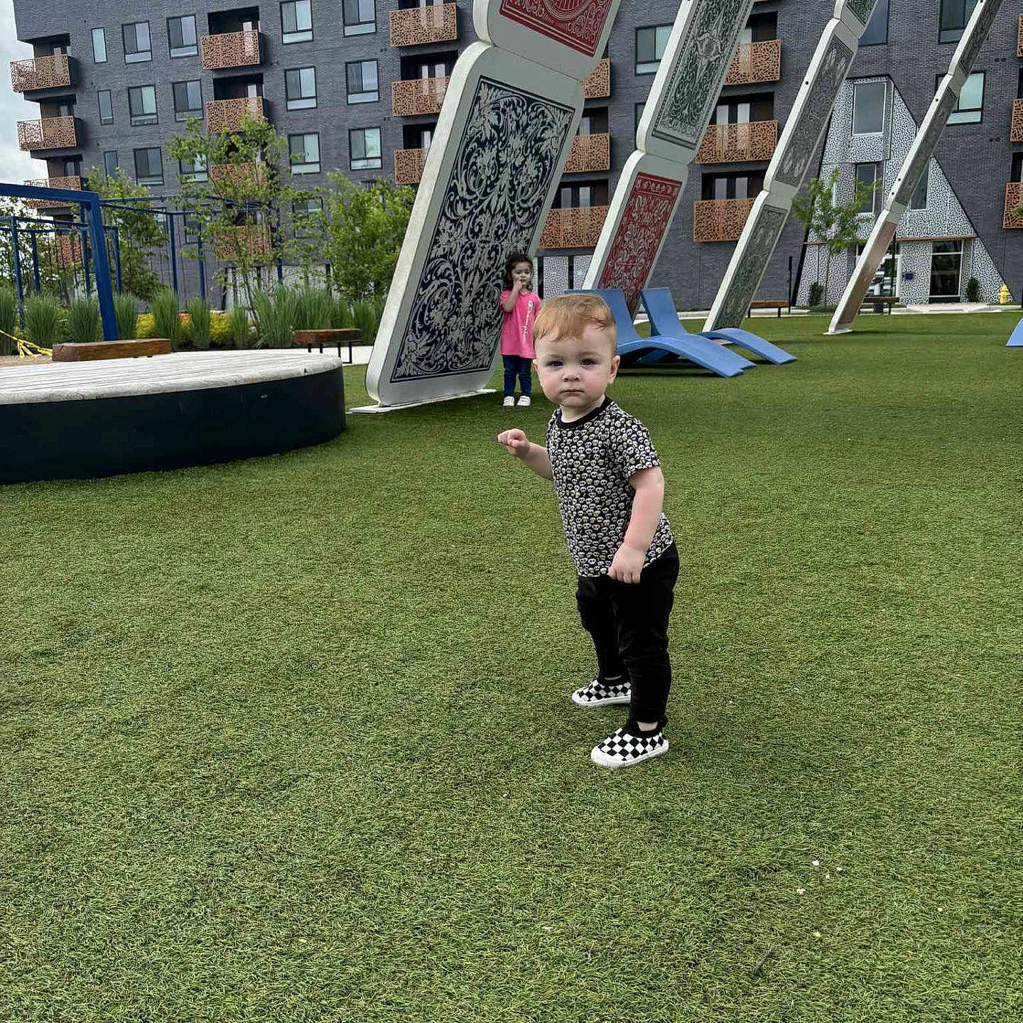 Celyn joined the competition — help win amazing prizes! architecture, building, casual_clothing, child, curious, daylight, fun, grass, greenery, modern, outdoor, park, person, play_area, playground, playing_cards, sneakers, toddler, urban, young_child