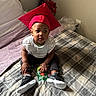 baby, bed, bedroom, black_pants, blanket, child, flowers, graduation_cap, happy, indoors, infant, lamp, photography, portrait, red_hat, shoes, sitting, snack_pouch, sneakers, white_shirt