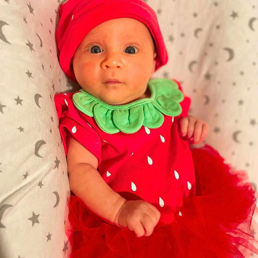 Delilah joined the competition — help win amazing prizes! baby, blanket, child, closeup, cute, festive, green_collar, hat, holiday_costume, indoors, infant, looking_at_camera, moon_pattern, person, portrait, red_clothing, smiling, stars_pattern, tulle_skirt, young_child