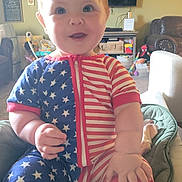 Ella is registered to the contest to win money with this photo: baby, blue, child, cushion, cute, face, furniture, happy, home, indoor, living_room, onesie, portrait, red, sitting, smiling, stars, stripes, toy, white