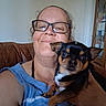 Chico is registered to the contest to win money with this photo: person, dog, glasses, smile, indoor, couch, brown, cabinet, small_dog, pet, holding, blue_shirt, face, fur, eyes, nose, ear, human, portrait, home