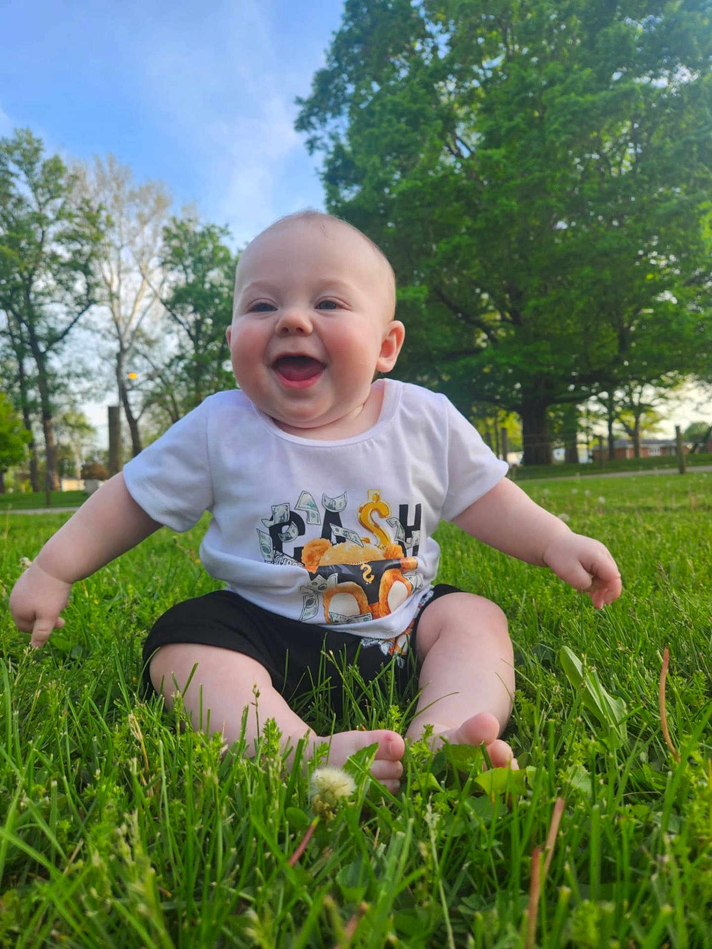 Ezra Ray is registered to the contest to win money with this photo: baby, blue_sky, child, cute, daytime, footwear_free, grass, greenery, happy, infant, nature, outdoor, park, playful, sitting, smiling, summer, sunlight, trees, tshirt
