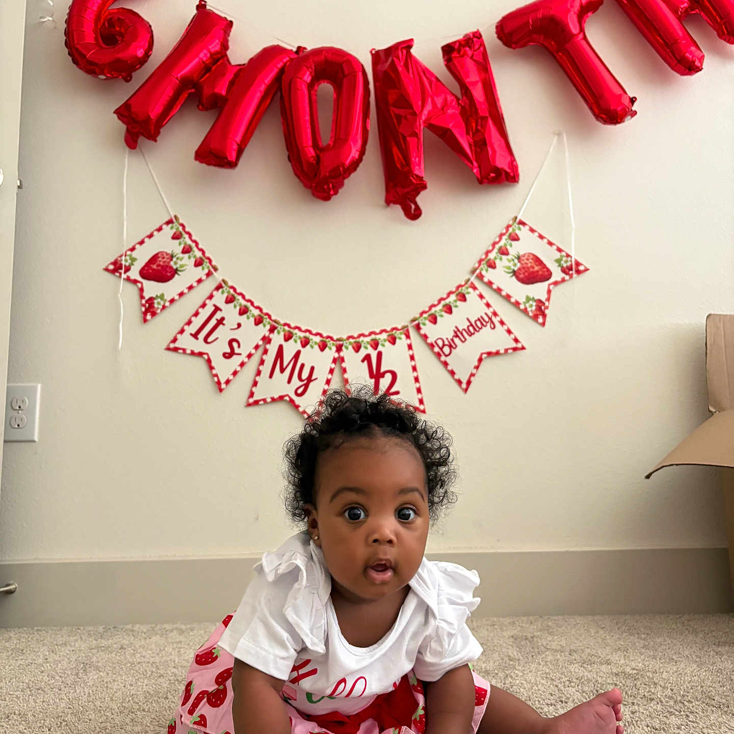 Rhyleigh is registered to the contest to win money with this photo: baby, child, infant, strawberry, red_balloon, birthday, half_birthday, carpet, white_wall, curly_hair, cute, sitting, decorations, celebration, earrings, clothing, indoor, portrait, smiling, young_child