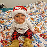 Raphaël participe au concours pour gagner de l'argent avec cette photo : baby, child, santa_hat, plaid_pants, yellow_booties, bedspread, floral_pattern, indoor, cute, holiday, christmas, smiling, sitting, person, apparel, warm_clothing, face, headwear, infant, cozy
