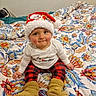 baby, child, santa_hat, plaid_pants, yellow_booties, bedspread, floral_pattern, indoor, cute, holiday, christmas, smiling, sitting, person, apparel, warm_clothing, face, headwear, infant, cozy