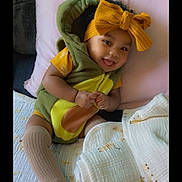 Rylaya is registered to the contest to win money with this photo: baby, child, smiling, yellow_bow, green_outfit, blanket, pillow, cute, indoors, sitting, happy, sock, furniture, portrait, cozy, person, headwear, infant, tongue_out, soft_toys