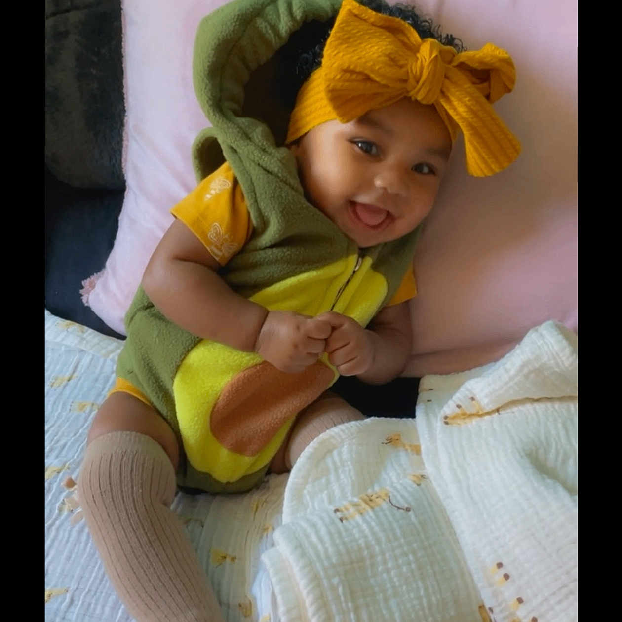 Rylaya is registered to the contest to win money with this photo: baby, child, smiling, yellow_bow, green_outfit, blanket, pillow, cute, indoors, sitting, happy, sock, furniture, portrait, cozy, person, headwear, infant, tongue_out, soft_toys