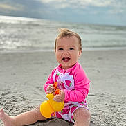 Catherine joined the competition — help win amazing prizes! baby, child, beach, sand, toy, rubber_duck, swimsuit, pink_clothing, strawberry_pattern, ocean, water, sky, clouds, happy, sitting, playful, smiling, barefoot, summer, outdoor