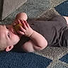 child, toddler, baby, lying_down, carpet, indoors, green_ball, toy, playing, face, eyes, hands, barefoot, gray_clothing, floor, home, portrait, casual, curious, happy