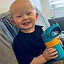 toddler, child, smiling, sitting, couch, sippy_cup, indoors, window, blinds, clothing, black_shirt, happy, light, face, hand, furniture, young_child, casual, home, portrait