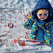 Abigaël a rejoint le concours — aidez-le/la à gagner de superbes lots ! baby, blanket, child, deer_illustration, fox_illustration, hood, milestone_blanket, numbers, outdoor, owl_illustration, portrait, ring, shadow, smile, snow, snowsuit, sunglasses, sunlight, toy, winter