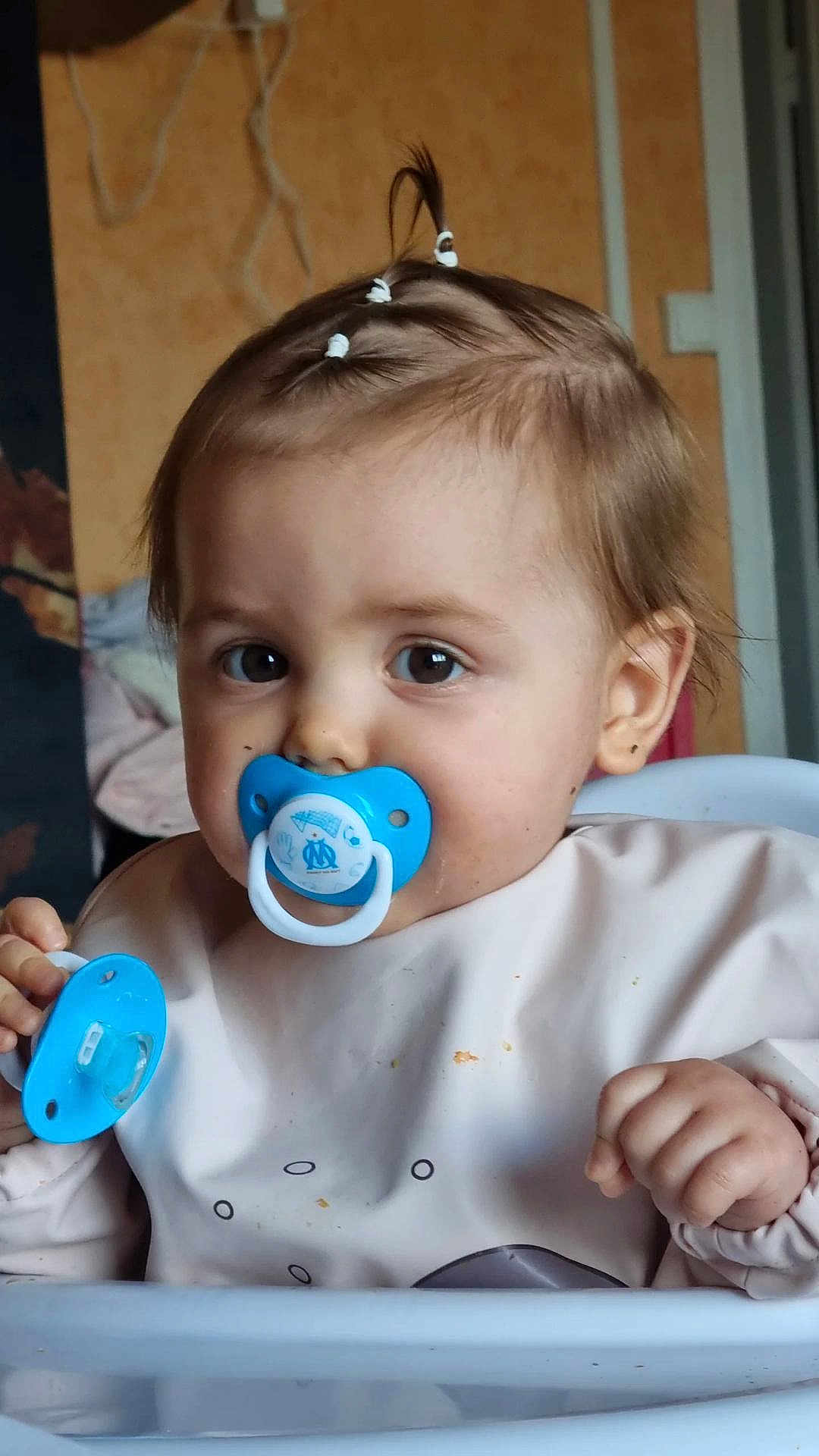Abigaël participe au concours pour gagner de l'argent avec cette photo : baby, bib, blue_pacifier, brown_eyes, child, close_up, clothing, cute, eyes, face, feeding, hair, hand, highchair, indoor, meal, pacifier, portrait, tiny_ponytail, toddler