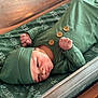 baby, newborn, infant, green_clothing, hat, blanket, fist, lying_down, bed, wooden_frame, face, skin, cute, sleepwear, portrait, small_hands, baby_bed, soft_fabric, peaceful, indoors