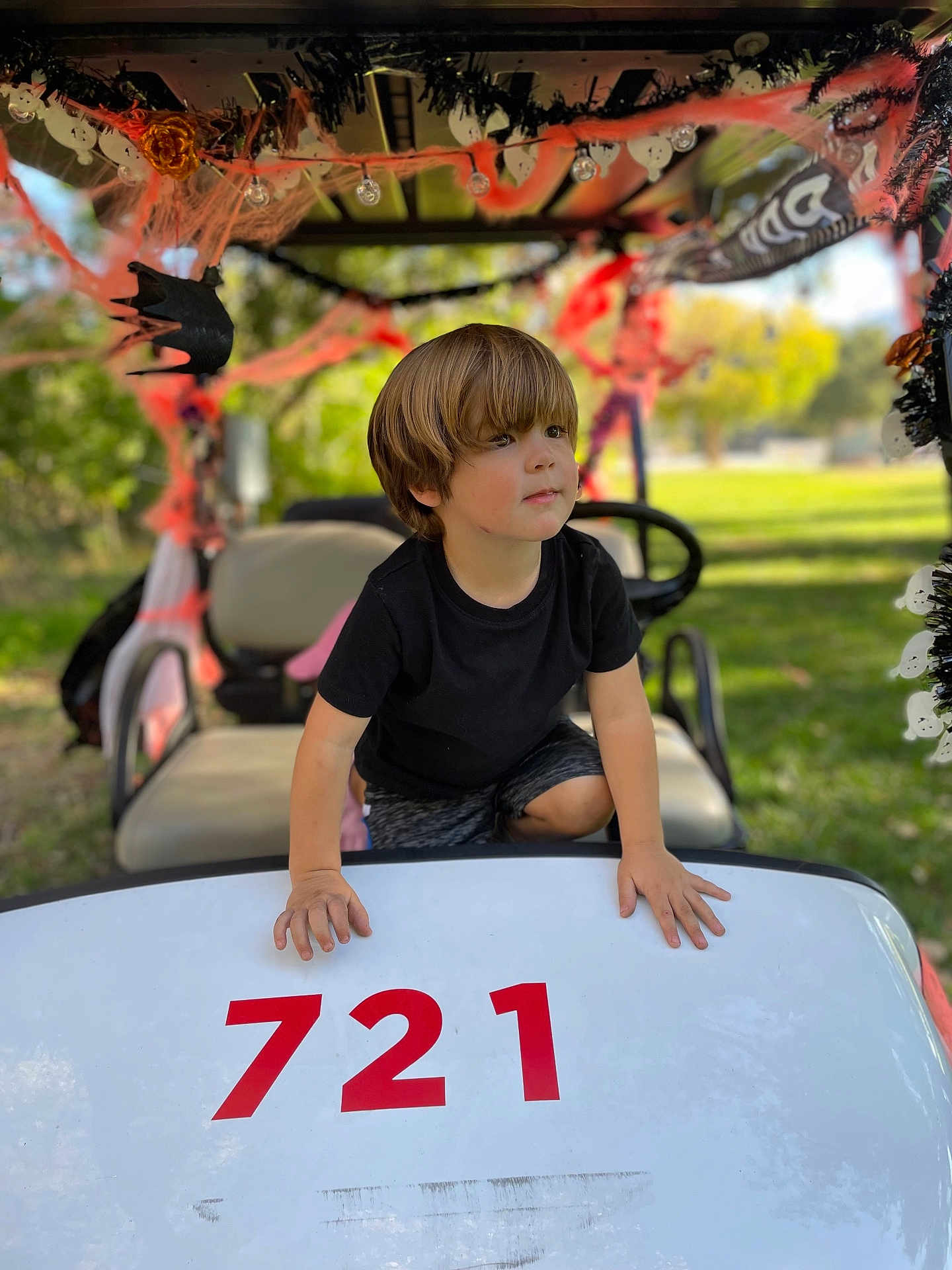 Mateo joined the competition — help win amazing prizes! child, boy, golf_cart, number, outdoor, daylight, halloween_decorations, spider_webs, orange, black, greenery, park, shorts, black_shirt, hands, face, hair, seat, steering_wheel, toy