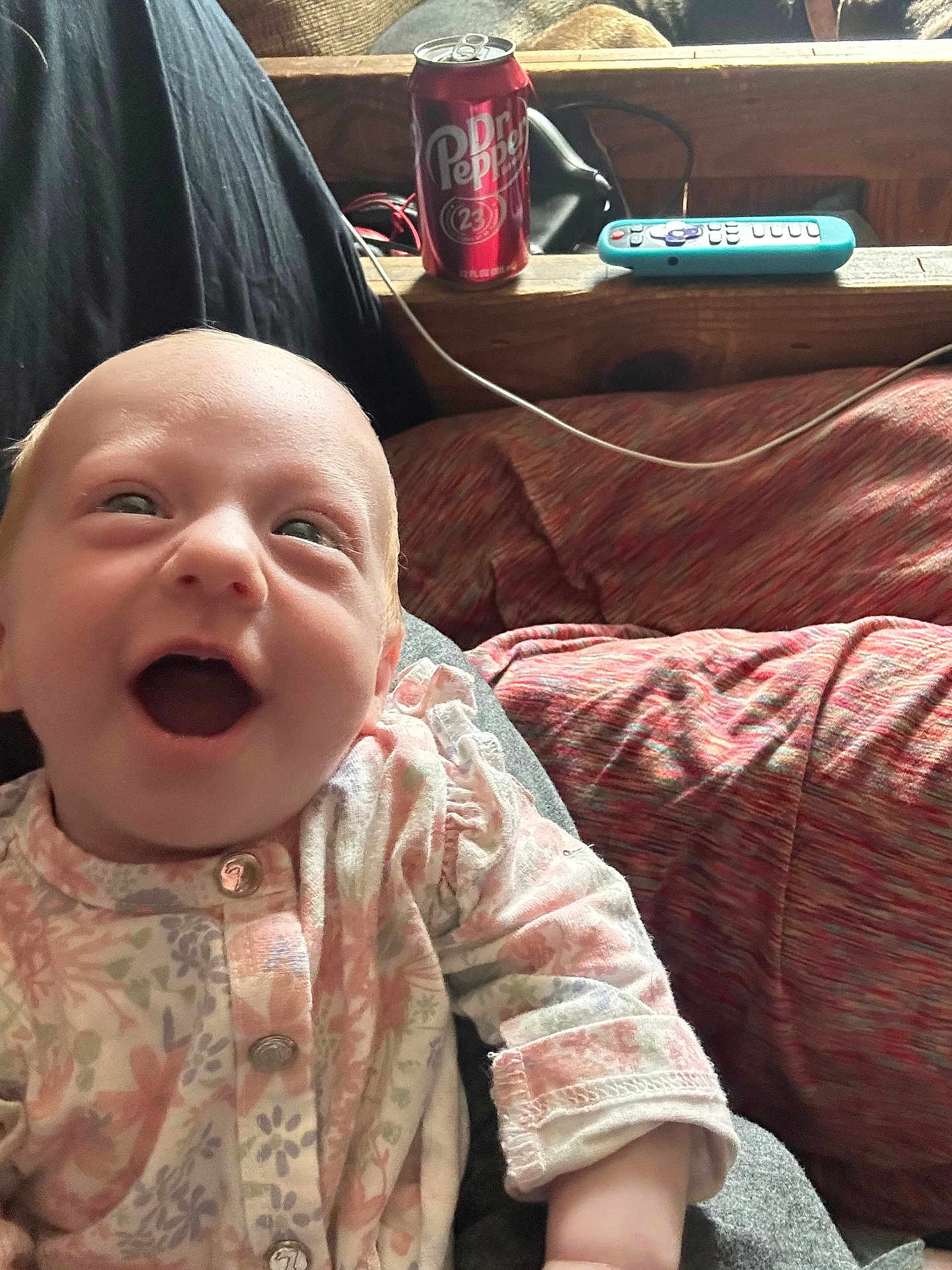 Tegan is registered to the contest to win money with this photo: baby, smiling, infant, onesie, floral_pattern, happy, face, indoor, couch, remote_control, beverage_can, wooden_surface, casual_clothing, child, person, cute, portrait, joyful, relaxed, home