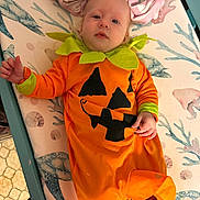 Tegan is registered to the contest to win money with this photo: baby, infant, costume, pumpkin, orange, green, face, lying_down, changing_pad, pattern, cute, child, holiday, halloween, clothing, indoor, person, looking, expression, hands