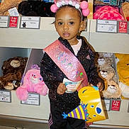 Lucia joined the competition — help win amazing prizes! toddler, birthday, sash, crown, party_hat, party_blower, stuffed_animals, toy_store, child, celebration, black_clothing, sneakers, floor_tiles, shelf, pink, yellow, smiling, indoor, young_child, portrait