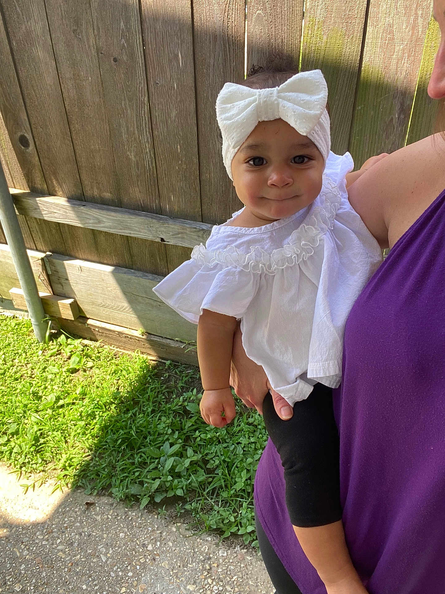Olivia joined the competition — help win amazing prizes! baby, child, headband, white_bow, white_top, black_leggings, person, adult, holding, outdoor, grass, wooden_fence, sunlight, shadow, side_view, smiling, cute, portrait, casual_clothing, greenery