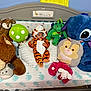 baby, tiger_costume, stuffed_animal, plush_toy, crib, bed, child, costume, animal_print, green_mushroom, deer, dragon, blue_elephant, pillow, pink_neck_pillow, toy, indoor, cute, sleeping_area, childrens_room