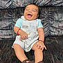 baby, laughing, smiling, overalls, sandals, couch, patterned_fabric, child, happy, seated, cute, infant, joyful, clothing, footwear, bracelet, portrait, indoors, expression, person