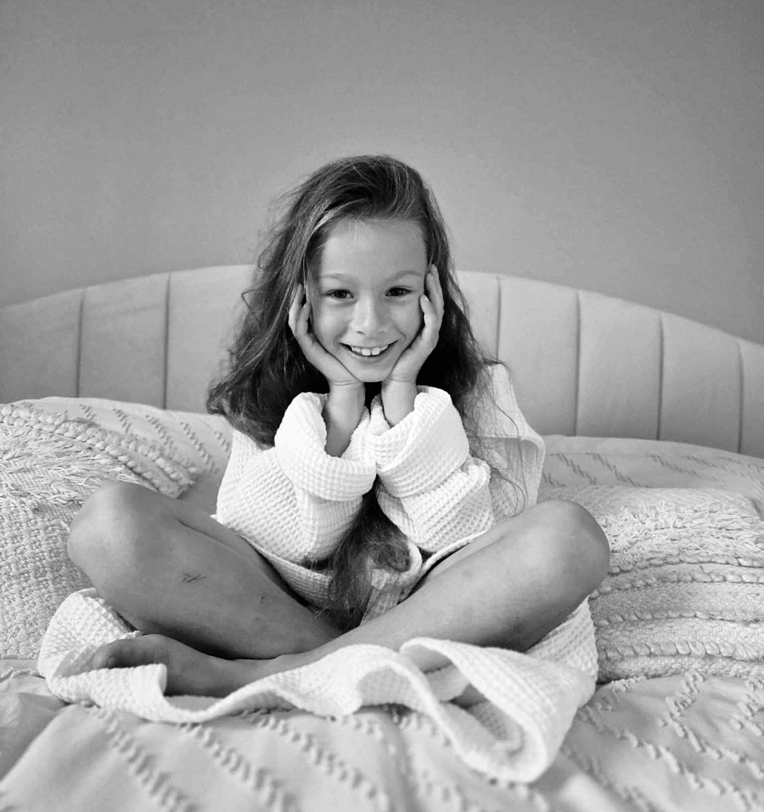 Lya participe au concours pour gagner de l'argent avec cette photo : bed, black_and_white, blanket, casual, child, comfortable, cozy, cross_legged, girl, happy, home, indoor, long_hair, monochrome, pillow, portrait, relaxed, robe, smiling, young
