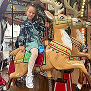 Lya a rejoint le concours — aidez-le/la à gagner de superbes lots ! amusement_park, background, carousel, child, clothing, decorative, entertainment, fun, girl, indoors, jacket, lights, playful, pole, reindeer, ride, smile, sneakers, tights, wooden_floor