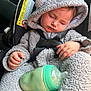 baby, sleeping, car_seat, bottle, hooded_outfit, gray_clothing, child, infant, safety_harness, peaceful, indoors, seat_belt, warning_label, hand, face, soft_texture, clothing, bottle_nipple, cute, resting