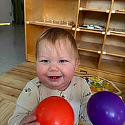 Lochlan is registered to the contest to win money with this photo: baby, child, smiling, happy, indoor, toy, ball, orange_ball, purple_ball, wooden_floor, wooden_shelf, play_area, caucasian_baby, long_sleeve_shirt, cute, portrait, toddler, face, hands, playtime