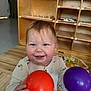 baby, child, smiling, happy, indoor, toy, ball, orange_ball, purple_ball, wooden_floor, wooden_shelf, play_area, caucasian_baby, long_sleeve_shirt, cute, portrait, toddler, face, hands, playtime