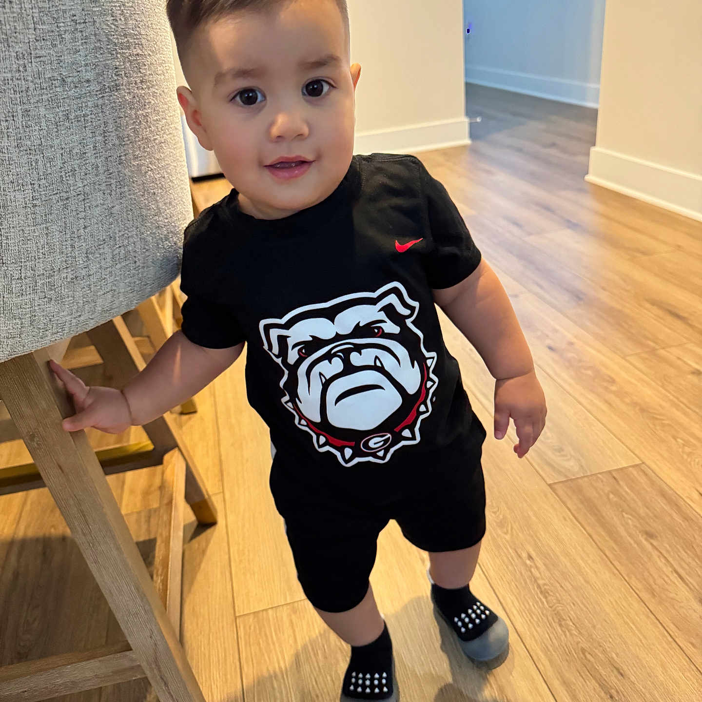 Manuell Carrillo joined the competition — help win amazing prizes! black_clothing, bulldog_logo, chair, child, curious_expression, face, floor, furniture, graphic_tshirt, hair, holding, home, indoor, shorts, slippers, smile, socks, standing, toddler, wooden_floor