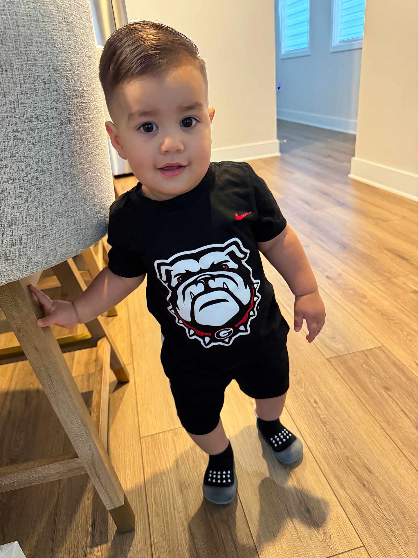Manuell Carrillo joined the competition — help win amazing prizes! black_clothing, bulldog_logo, chair, child, curious_expression, face, floor, furniture, graphic_tshirt, hair, holding, home, indoor, shorts, slippers, smile, socks, standing, toddler, wooden_floor