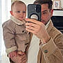 Elyo participe au concours pour gagner de l'argent avec cette photo : baby, man, selfie, mirror, smartphone, corduroy_jacket, beige_clothing, holding, portrait, indoor, living_room, parent, child, toddler, hand, face, smile, reflection, window_light, casual