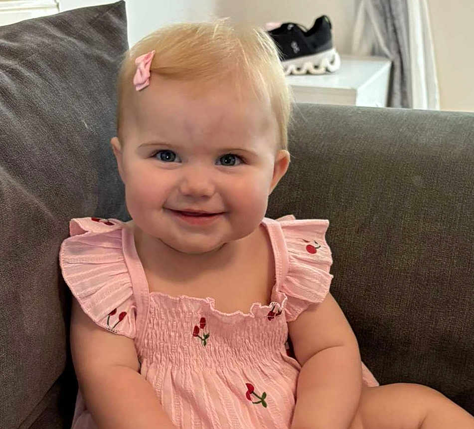 Isla-Rose is registered to the contest to win money with this photo: baby, child, smile, pink_dress, bow, couch, indoor, face, happy, arm, sitting, furniture, blond_hair, casual, cute, young_child, person, footwear, background, comfortable