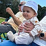 adult, baby, blue_pants, casual, child, daylight, family, grass, hand, happy, hat, lap, nature, outdoor, person, sitting, smile, sunglasses, watch, white_clothing