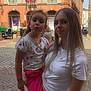 Mia participe au concours pour gagner de l'argent avec cette photo : child, teenager, outdoor, street, cobblestone, building, window, flag, person, girl, smile, pose, white_shirt, pink_pants, bracelet, necklace, daylight, sky, urban, historic_building