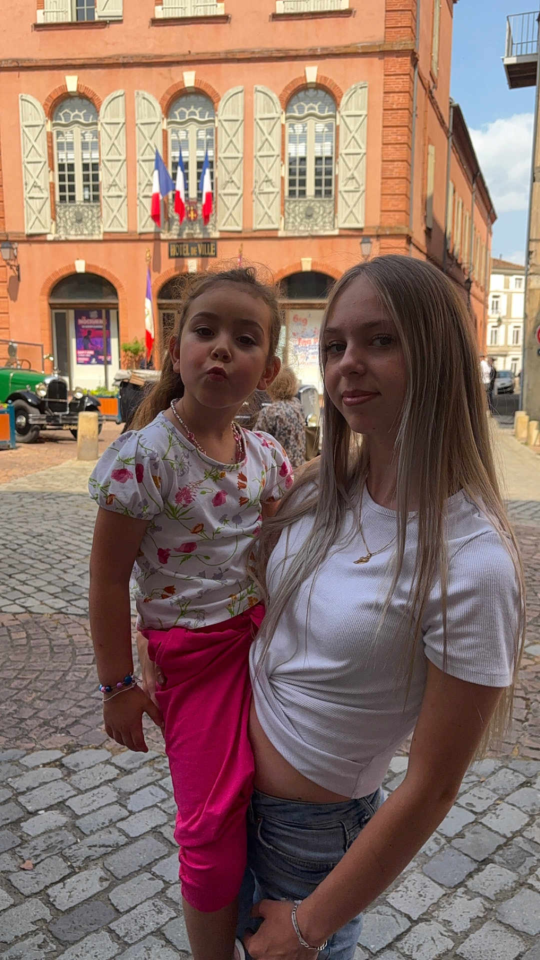 Mia participe au concours pour gagner de l'argent avec cette photo : child, teenager, outdoor, street, cobblestone, building, window, flag, person, girl, smile, pose, white_shirt, pink_pants, bracelet, necklace, daylight, sky, urban, historic_building