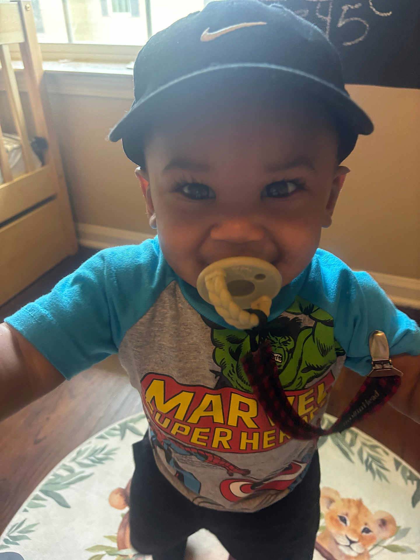 Kyrian joined the competition — help win amazing prizes! toddler, child, pacifier, marvel, superhero, shirt, cap, nike, smiling, indoors, rug, animal_print, floor, wooden_floor, window, baby, cute, happy, person, clothing