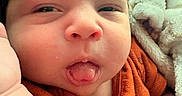 Stiles is registered to the contest to win money with this photo: baby, blanket, child, close_up, cozy, cute, expression, face, hand, indoors, infant, newborn, orange_clothing, person, portrait, skin, sleepy, soft_texture, tongue, warm_colors