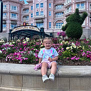 Ylona participe au concours pour gagner de l'argent avec cette photo : toddler, child, flower_bed, plush_toy, pink_shorts, white_tshirt, sneakers, disneyland_park, clock_tower, topiary, building, balcony, blue_sky, clouds, flowering_plants, stone_ledge, outdoor, smiling, daytime, tourist_attraction