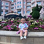 toddler, child, flower_bed, plush_toy, pink_shorts, white_tshirt, sneakers, disneyland_park, clock_tower, topiary, building, balcony, blue_sky, clouds, flowering_plants, stone_ledge, outdoor, smiling, daytime, tourist_attraction