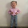 toddler, child, smiling, standing, pigtails, pink_shirt, jeans, sneakers, indoor, wooden_floor, plain_wall, happy, casual_clothing, small_person, cute, portrait, fashion, young_child, full_body, playful