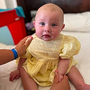 Evie is registered to the contest to win money with this photo: baby, child, infant, yellow_dress, sitting, bed, hands, bracelet, manicure, blue_eyes, soft_focus, indoor, person, face, cute, skin, headboard, furniture, red_object, blue_object
