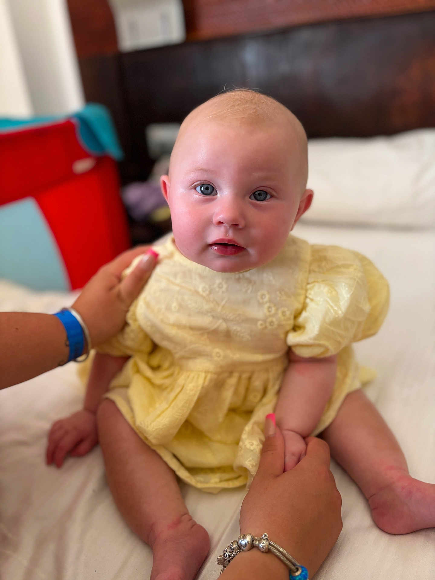 Evie is registered to the contest to win money with this photo: baby, child, infant, yellow_dress, sitting, bed, hands, bracelet, manicure, blue_eyes, soft_focus, indoor, person, face, cute, skin, headboard, furniture, red_object, blue_object