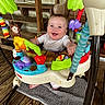 baby, smiling, activity_jumper, toy, colorful, happy, infant, wooden_floor, bench, indoor, child, playtime, face, person, seat, floor_mat, decor, purple_toy, red_toy, green_toy
